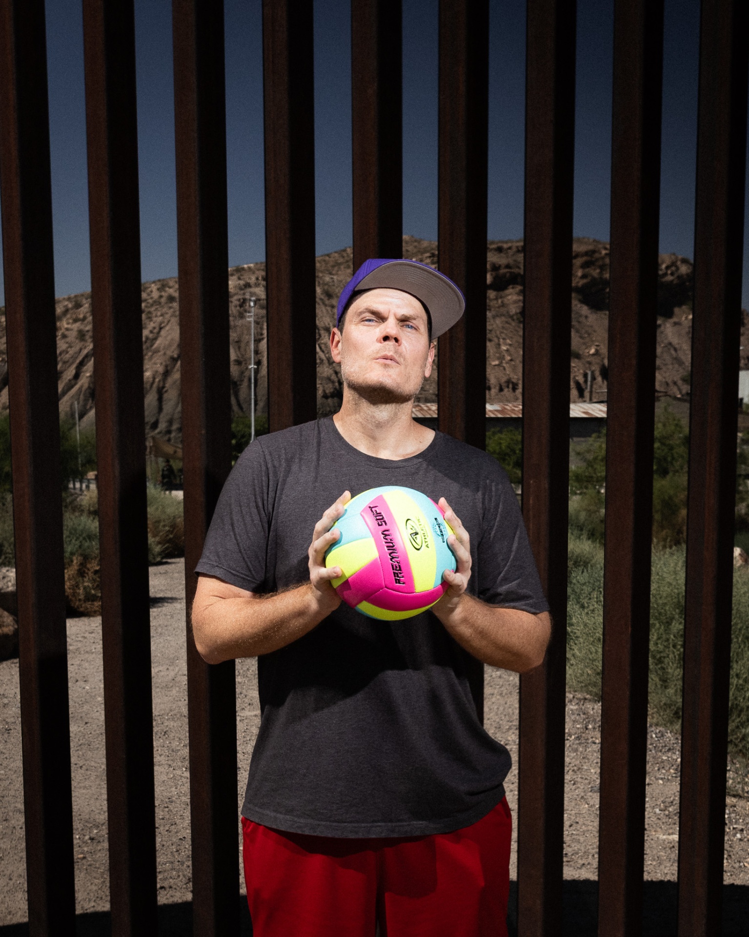 Man in a backwards cap holding a volleyball at the border fence