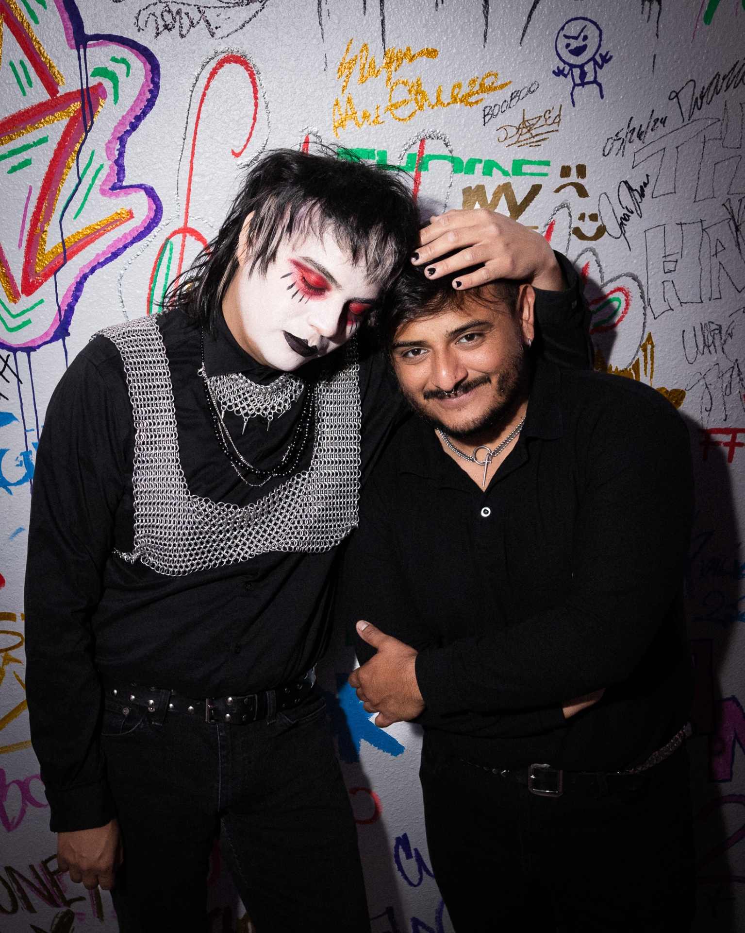 Two people against a graffiti-covered wall, one in dramatic goth makeup